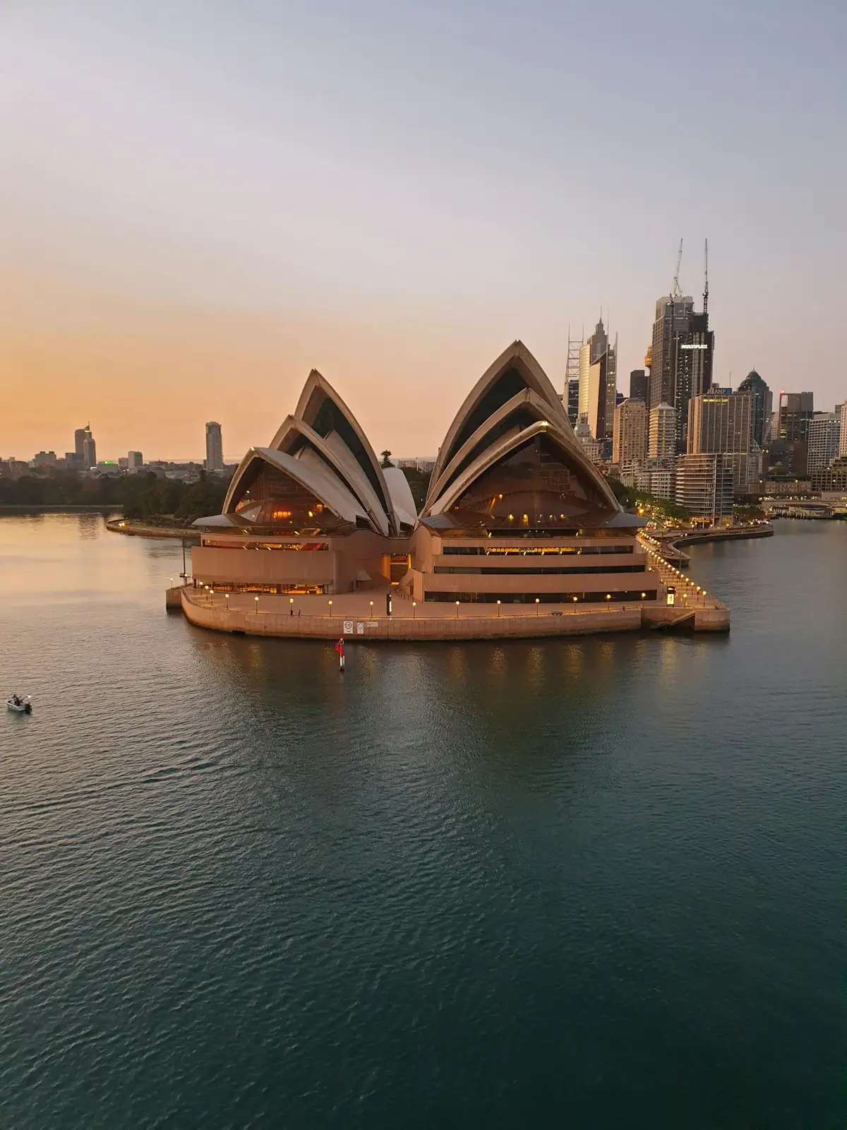 The Sydney Opera House on Bennelong Point with the city skyline behind, under a peach sunrise sky over still harbour water
