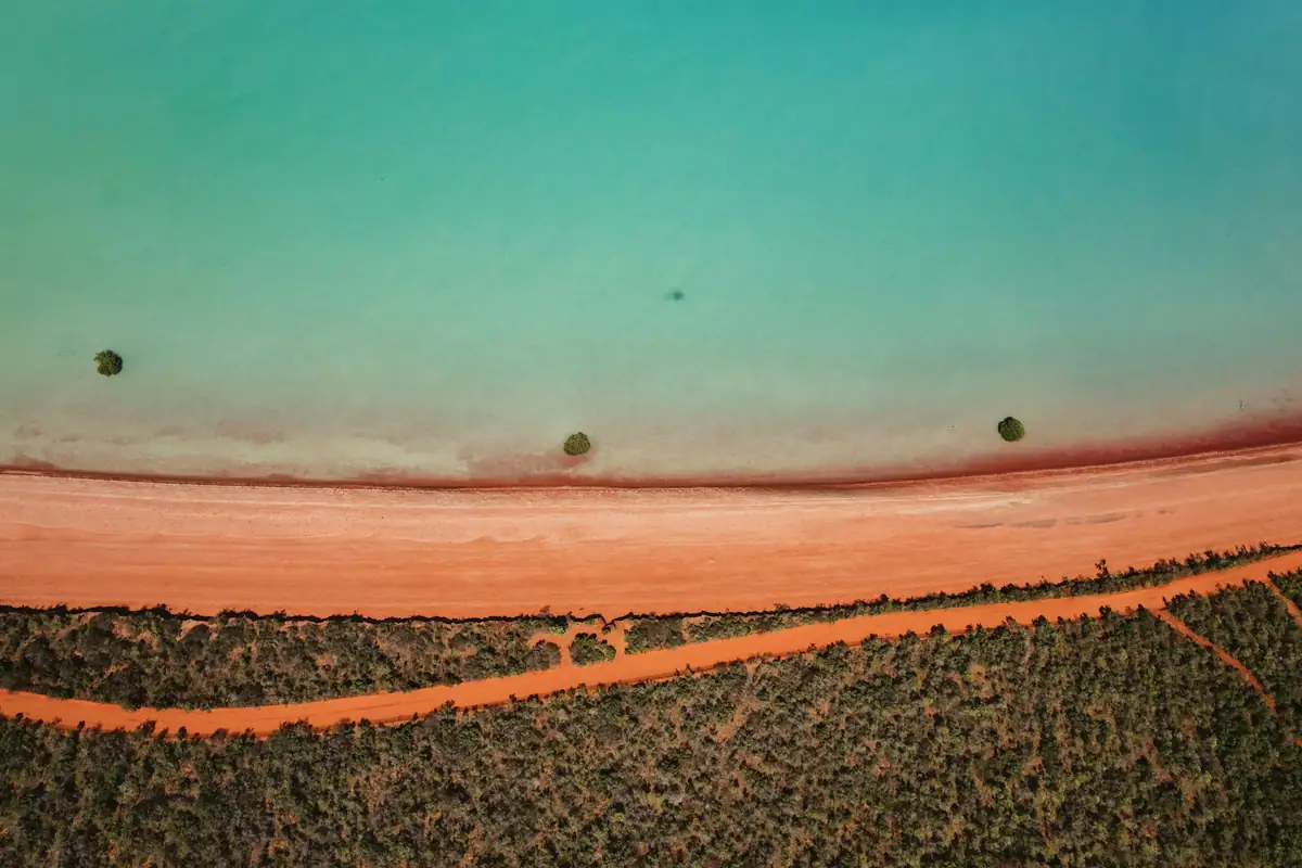 Top-down aerial of turquoise sea meeting a strip of red-orange sand and green scrub along the Broome coast
