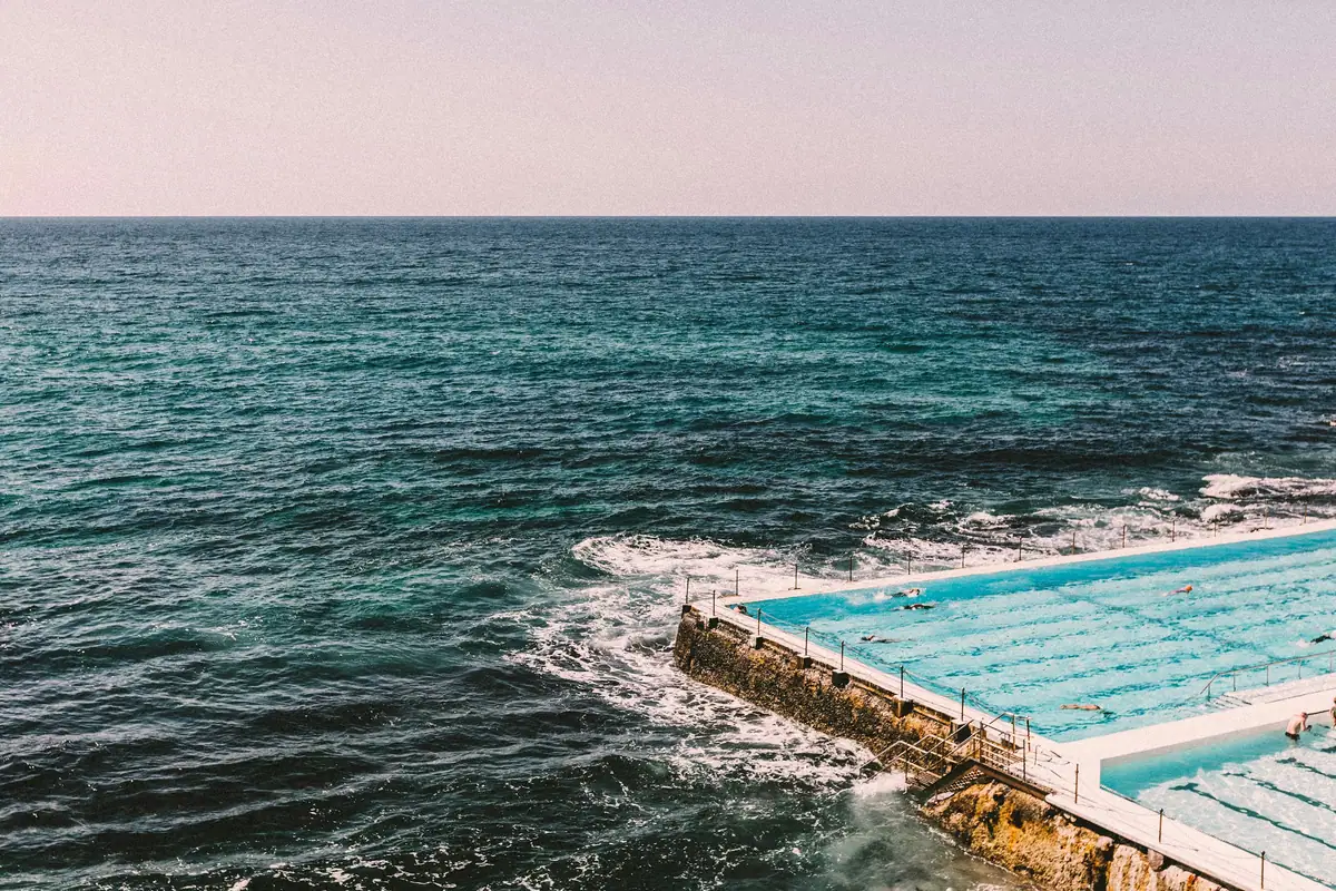 Waves wash over the edge of a turquoise ocean pool on the rocks at Bondi, with open sea to the horizon