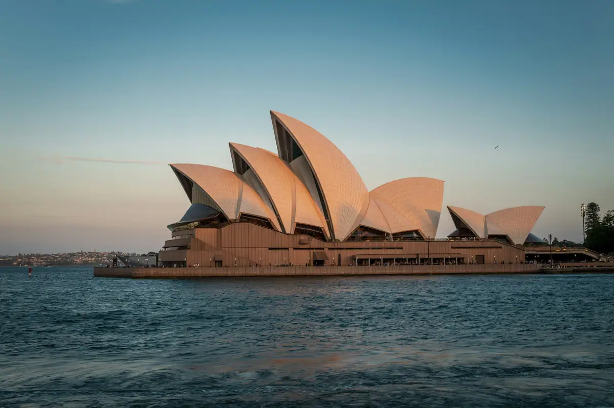 The Sydney Opera House sails glow pink in evening light across choppy harbour water
