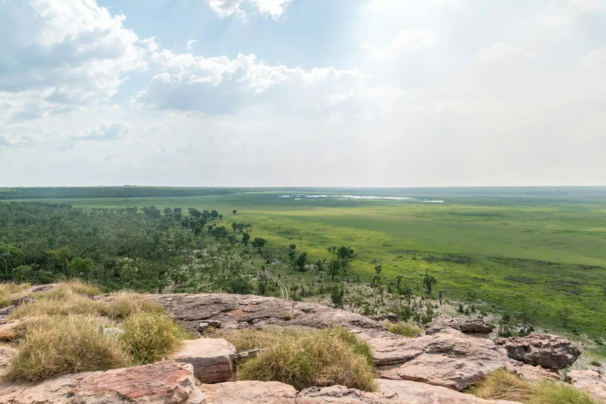 From a rocky sandstone lookout, bright green wetland floodplains stretch to the horizon beneath a cloudy sky in Kakadu